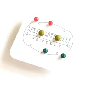 Pode incluir: Um conjunto de seis brincos de pressão em três cores: verde, amarelo e rosa. Os brincos são exibidos em um cartão branco com o texto "Leet Love Dale Jewelry".