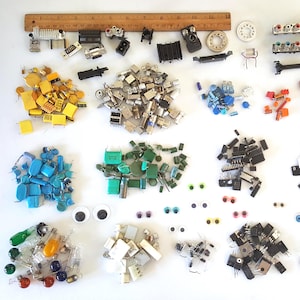 May include: A collection of various electronic components, including capacitors, resistors, and integrated circuits, arranged on a white surface. The components are in various colors, shapes, and sizes, with a wooden ruler at the top. A variety of keyboard keys and googly eyes are also present.