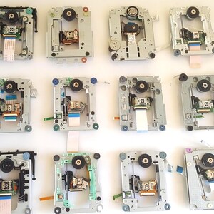 May include: Twelve silver and metallic optical disc drive laser lens mechanisms arranged in a grid. Each unit features a central motor, a sliding optical pickup head, circuit boards, and flexible ribbon cables for data connection.
