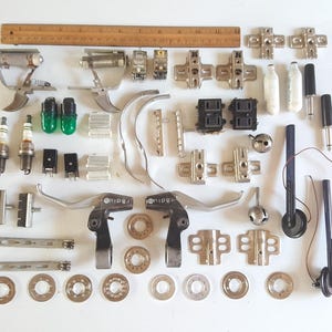 May include: An assortment of small metal and plastic parts, including hinges, levers, and electrical components, arranged on a white surface. A wooden ruler is at the top. The parts vary in size and shape, with silver, black, and green elements.
