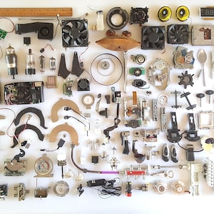 May include: A collection of assorted electronic components, mechanical parts, and hardware arranged on a white surface, including cooling fans, vacuum tubes, circuit boards, a wooden ruler measuring 12 inches, and various metal gears and connectors.