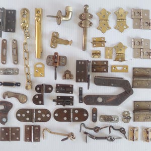 May include: A collection of various metal hardware pieces, including hinges, latches, and door stops. The hardware is in various finishes, including brass, bronze, and silver. The items are arranged on a white surface.