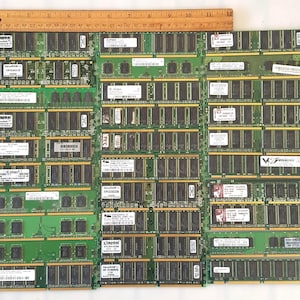 May include: A collection of green computer RAM modules with gold connectors, various labels, and a wooden ruler. The modules are arranged in a grid, showcasing their components and design. The ruler is approximately 12 inches long.