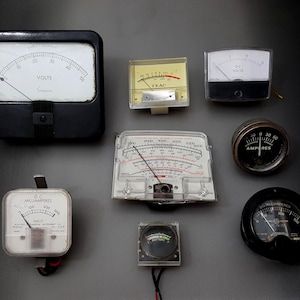 May include: A collection of eight vintage analog electrical meters and gauges, including various rectangular and circular designs with black and white faces, needle indicators, and printed scales for measuring volts, amperes, and battery levels.