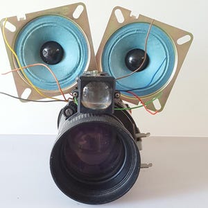 May include: A unique assemblage featuring two light blue speakers with black centers, positioned above a camera lens. The speakers are mounted on square metal frames. A clear lens and various wires complete the design, creating a mechanical art piece.
