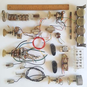 May include: A collection of vintage electronic components including metal variable capacitors, transformers, potentiometers with knobs, wiring, small light bulbs, and a rectangular control panel, all arranged near a 12-inch wooden ruler for scale.