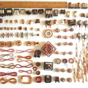 May include: An assortment of electronic components, including coils, transformers, and wire, arranged on a white surface. A wooden ruler is at the top. The components vary in size, shape, and color, with copper wire and red and brown components.