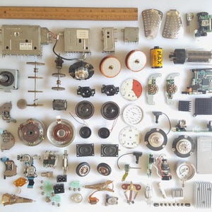 May include: A collection of assorted electronic components and mechanical parts arranged on a white surface, including circuit boards, small speakers, dials, gears, a 35mm film canister, and a 12-inch wooden ruler for scale.