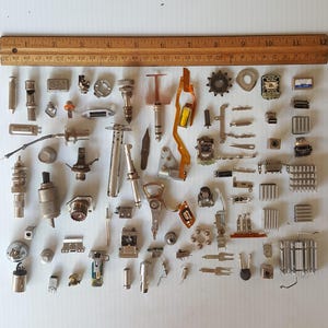 May include: A collection of small, metallic electronic components and hardware, including connectors, gears, and circuit board parts. A wooden ruler with inch markings is at the top of the image. The components are arranged on a white surface.