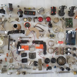 May include: A collection of various electronic components, including circuit boards, speakers, dials, and metal parts, arranged on a white surface. The components are in various shapes and sizes, with a mix of colors like black, silver, orange, and red.