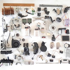 May include: An assortment of electronic components, including speakers, motors, and circuit boards, are arranged on a white surface. Various sizes and shapes are visible, with a wooden ruler at the top. The components are in shades of black, gray, brown, and white.
