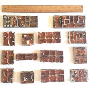 May include: A collection of thirteen rectangular electronic circuit boards with brown substrates and silver metal shielding partitions, arranged in rows. A wooden ruler measuring 12 inches is positioned horizontally above the components.