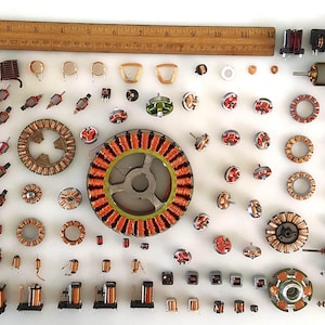 May include: A collection of various electrical components, including coils, armatures, and rotors, arranged on a white surface. A wooden ruler, approximately 12 inches long, is placed at the top. The components feature copper wiring and metal parts.