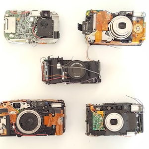 May include: Five disassembled digital cameras with exposed circuit boards, lenses, and internal components. The cameras are arranged in a grid pattern on a white surface.