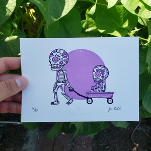May include: A purple and white print of two skeleton figures with floral skull designs. The larger skeleton is pulling a wagon with the smaller skeleton inside. The wagon has a teddy bear in it. The print is numbered 79/85 and signed "Joan Palada".