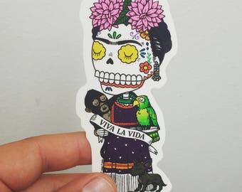 Puede incluir: Una pegatina con una ilustración de dibujos animados de Frida Kahlo con una cara de calavera de azúcar, usando una corona de flores y un vestido tradicional mexicano. El texto "Viva la Vida" está escrito en una pancarta en su mano. Ella está sosteniendo un mono y un loro.