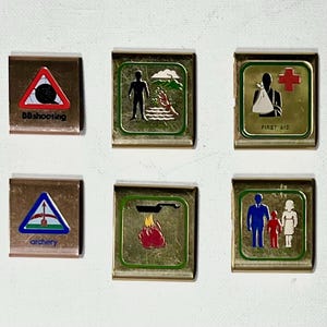 May include: A collection of vintage metal pins with various activity and service emblems. The pins feature images and text for activities like BB shooting, archery, first aid, and running. The pins have a gold border and are set against a white background.