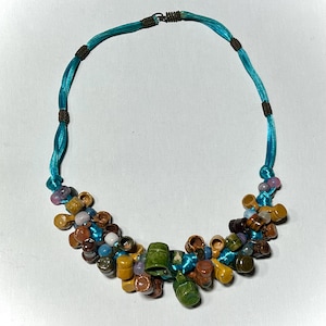 May include: A turquoise blue ribbon necklace with a cluster of colorful ceramic beads. The beads are in various shapes and sizes, including small pots, spheres, and cylinders. The beads are in shades of brown, green, yellow, blue, and purple.