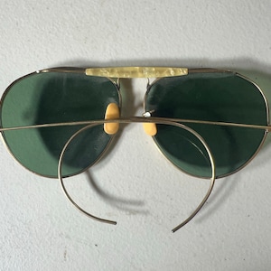 Op de afbeelding: Gouden aviator zonnebril met groene glazen en een neusbrug. De zonnebril heeft een draadframe en is dubbelgevouwen.
