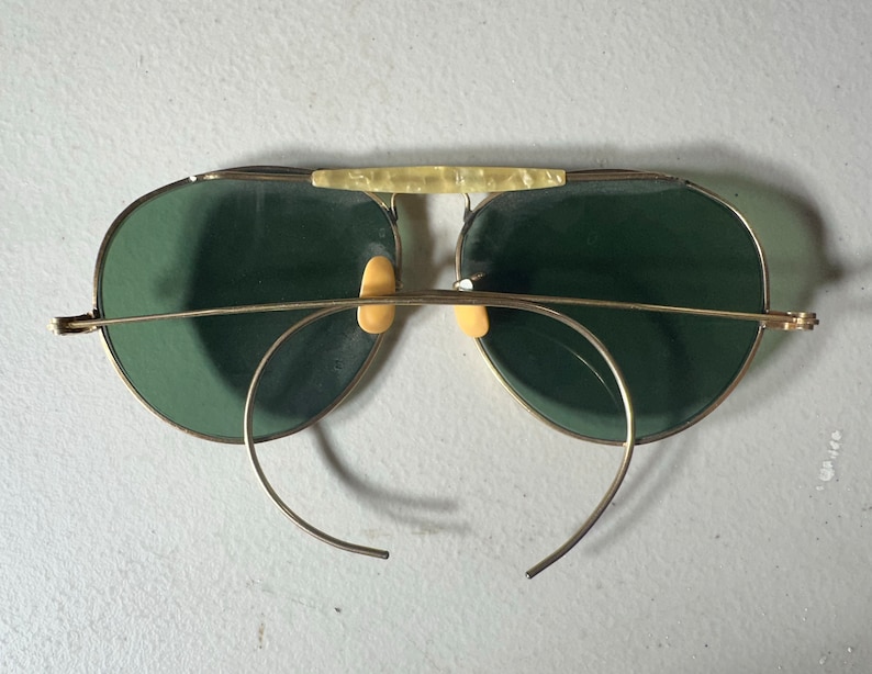 Op de afbeelding: Gouden aviator zonnebril met groene glazen en een neusbrug. De zonnebril heeft een draadframe en is dubbelgevouwen.