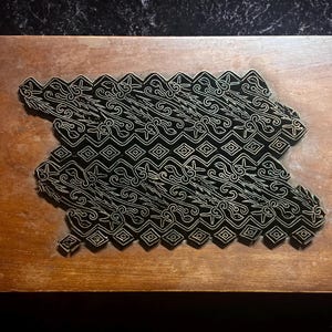 May include: A wooden block with a carved design, used for printing patterns onto fabric. The design is intricate and features a repeating pattern of swirls and diamonds.