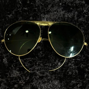 Op de afbeelding: Gouden aviator zonnebril met donkergroene glazen. De zonnebril ligt op een zwarte achtergrond.