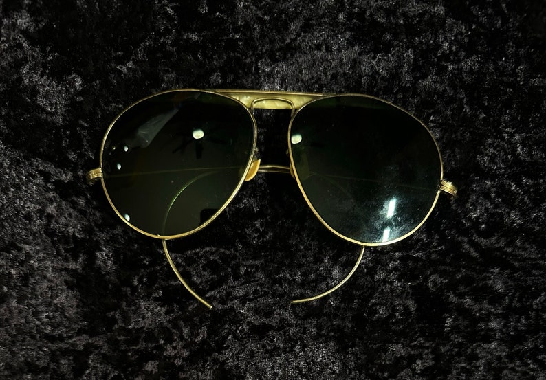 Op de afbeelding: Gouden aviator zonnebril met donkergroene glazen. De zonnebril ligt op een zwarte achtergrond.