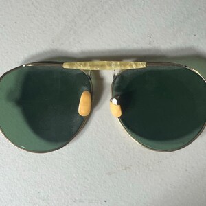Op de afbeelding: Een paar gouden aviator zonnebrillen met groene glazen en een cr&egrave;mekleurige brug.