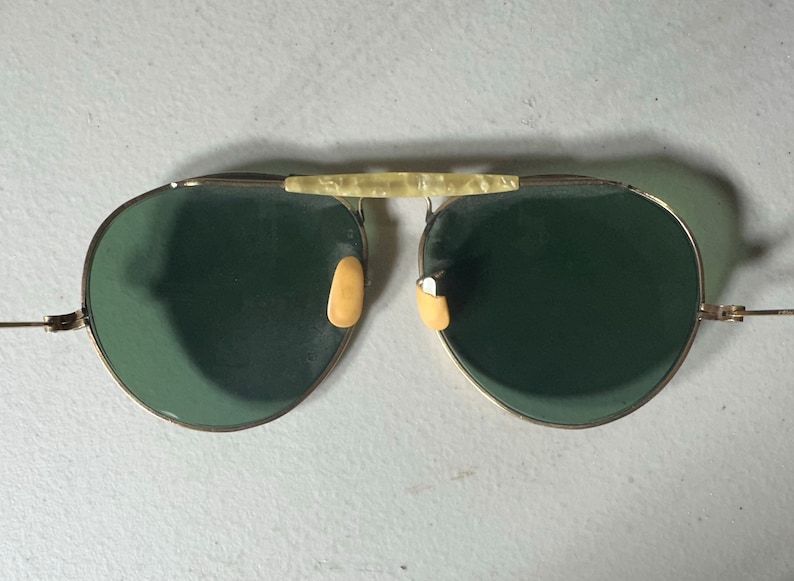 Op de afbeelding: Een paar gouden aviator zonnebrillen met groene glazen en een cr&egrave;mekleurige brug.