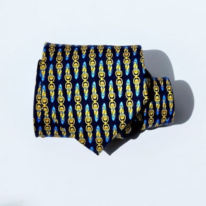 May include: A folded navy blue tie with a repeating pattern of gold and light blue chain-like links. The tie is made of a smooth fabric and is displayed on a white surface. The tie is a classic accessory for formal or business attire.
