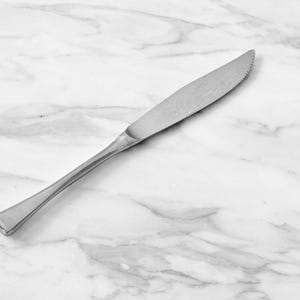May include: A stainless steel butter knife with a serrated edge on a white marble surface. The knife has a long, slender handle and a slightly curved blade, ideal for spreading butter or other soft foods.