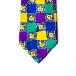 May include: A patterned necktie with a repeating checkerboard design. The squares alternate between denim blue, yellow, green, purple, and blue. Each square contains a gold floral design. The tie is on a white background.
