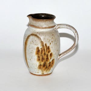 May include: A handmade ceramic pitcher with a speckled, off-white glaze. The pitcher features a brown rim and handle, with a decorative brown pattern on the front. The pitcher has a capacity of approximately 48 ounces.