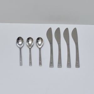 May include: A set of seven stainless steel utensils consisting of three spoons and four butter knives. Each piece features a smooth, polished finish and a small, distinct engraved emblem located near the base of the handle on the front side.