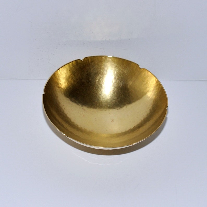 Puede incluir: Un cuenco de metal de color dorado con un borde festoneado. El cuenco es liso y brillante, y tiene una textura ligeramente martillada.