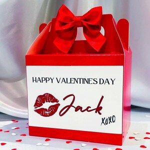 May include: Red gift box with a red bow on top. The box has a white label with the text "Happy Valentines Day" and a red lipstick print. The name "Jack" is written in cursive on the label with the text "XOXO" below it.