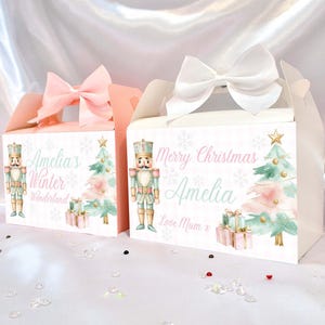 Può includere: Due scatole regalo con fiocchi bianchi. Una scatola è rosa, l'altra bianca. Entrambe le scatole presentano un'illustrazione di schiaccianoci, un albero di Natale e il testo "Amelia's Winter Wonderland" e "Merry Christmas Amelia".