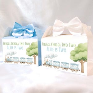 Könnte beinhalten: Zwei Geschenkboxen, eine blaue und eine weiße, mit Schleifenverschlüssen. Jede Box zeigt eine Zugillustration, einen Baum und den Text "CHUGGA CHUGGA TWO TWO, ALFIE IS TWO".