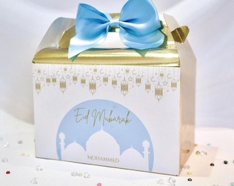 May include: A gold gift box with a blue bow and a white label that says "Eid Mubarak" and "Mohammed". The box is decorated with a pattern of crescent moons and lanterns.