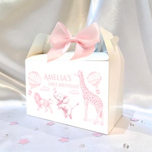 May include: A white party favor box with a pink bow on top. The box is decorated with pink illustrations of a giraffe, lion, elephant, and hot air balloons. The text "AMELIA'S FIRST BIRTHDAY" is printed on the front.