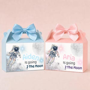 May include: Two rectangular cardboard favor boxes with handles and satin bows. The left box is light blue with an astronaut graphic and the text "Aiden Is going 2 the Moon". The right box is light pink with an astronaut and "Aria Is going 2 the Moon".