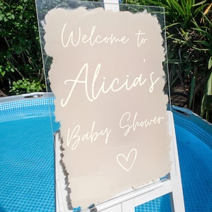 May include: A clear acrylic sign with a beige background and white lettering. The sign says "Welcome to Alicia's Baby Shower" with a white heart at the bottom.