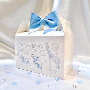 Può includere: Scatola regalo bianca con un fiocco blu in cima. La scatola ha un design blu e bianco con una giraffa, un elefante e un leone. Il testo sulla scatola dice "Reuben's First Birthday."