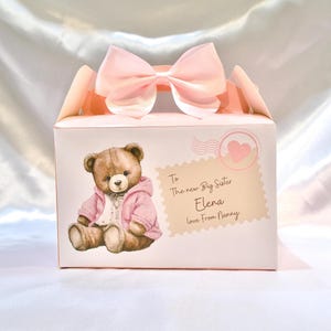 Op de afbeelding: Roze cadeaudoos met een roze strik en een teddybeerillustratie. De doos heeft een bericht met de tekst "Voor de nieuwe grote zus Elena Liefs van oma".