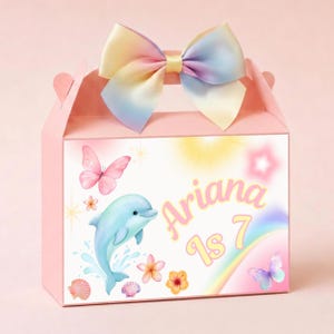 Könnte beinhalten: Rosa Party-Geschenkbox mit einer pastellfarbenen Regenbogen-Schleife. Die Box zeigt einen Delfin, Schmetterlinge, Blumen und den Text "Ariana is 7". Das Design umfasst einen Regenbogen und Muscheln.