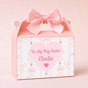May include: A pink gift box with a large pink bow on top. The box features a heart-shaped design with the text "To My Big Sister Elodie". The box is decorated with bows and hearts.