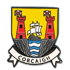 May include: A yellow and red enamel pin with a coat of arms design. The coat of arms features two red towers with white crosses on top, a ship with white sails, and a blue and white wavy line. The bottom of the pin has a white banner with the text "CORCAIGH" in black.
