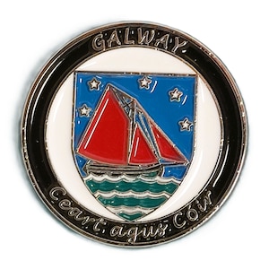 May include: A silver-colored metal pin with a black and white circular design. The center features a red sailboat with a blue and white background. The text "GALWAY" is written in a black circle around the sailboat. The text "Ceart agus Cóir" is written in a black circle around the bottom of the sailboat.
