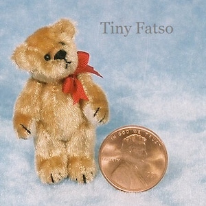 Puede incluir: Un osito de peluche en miniatura con pelaje marrón claro, ojos negros y un lazo rojo. El oso está junto a una moneda para comparar el tamaño. El texto "Tiny Fatso" está en el fondo.