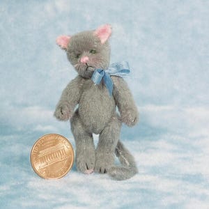 May include: A small gray plush cat with a blue bow, standing next to a US penny.
